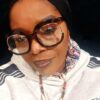 Close-up portrait of a woman wearing oversized round glasses, a head scarf, and a beaded necklace.