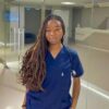 Young woman in blue scrubs with a stethoscope, standing in a glass-walled hospital hallway.