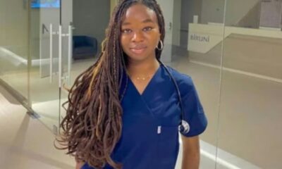 Young woman in blue scrubs with a stethoscope, standing in a glass-walled hospital hallway.