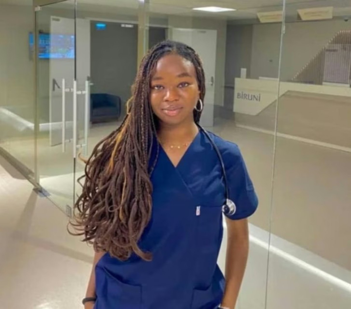 Young woman in blue scrubs with a stethoscope, standing in a glass-walled hospital hallway.