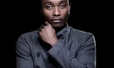 Portrait of a man with his hand near his chin, wearing a gray turtleneck sweater, against a black background.
