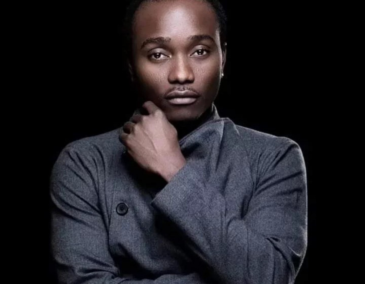 Portrait of a man with his hand near his chin, wearing a gray turtleneck sweater, against a black background.