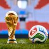 Gold World Cup trophy standing on grass beside a colorful soccer ball on a stadium field.