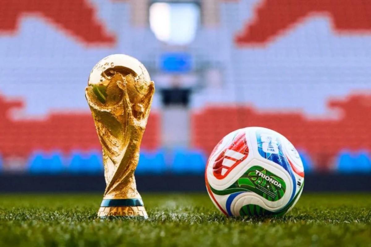 Gold World Cup trophy standing on grass beside a colorful soccer ball on a stadium field.