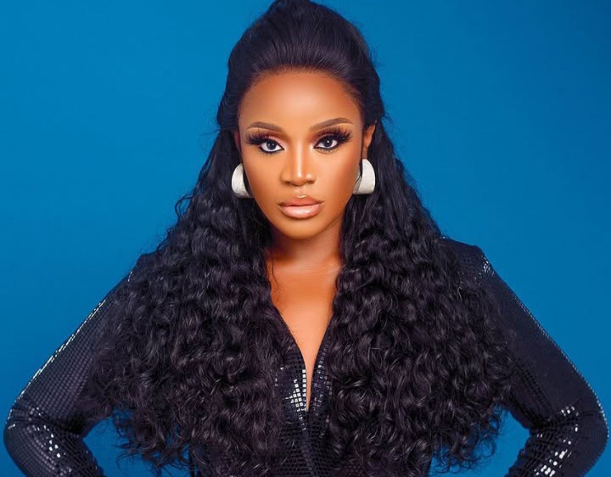 Portrait of a woman with long, curly black hair, bold makeup, white statement earrings, and a black mesh outfit against a blue background.