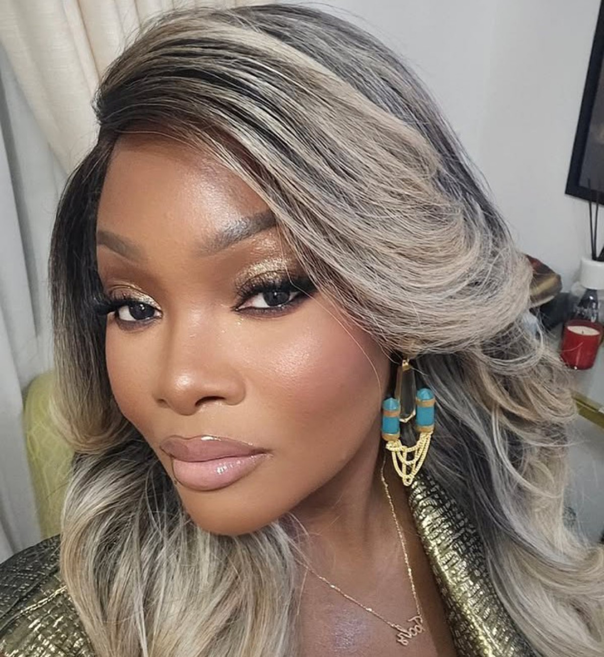Close-up of a woman with ash-blonde hair, golden makeup, and turquoise gold earrings, looking at the camera.