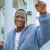 Smiling man in a light blue fleece jacket raises a clenched fist in the air outdoors, pointing with his other hand in a celebratory pose.