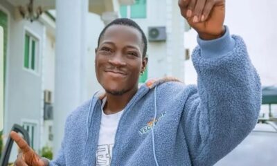 Smiling man in a light blue fleece jacket raises a clenched fist in the air outdoors, pointing with his other hand in a celebratory pose.