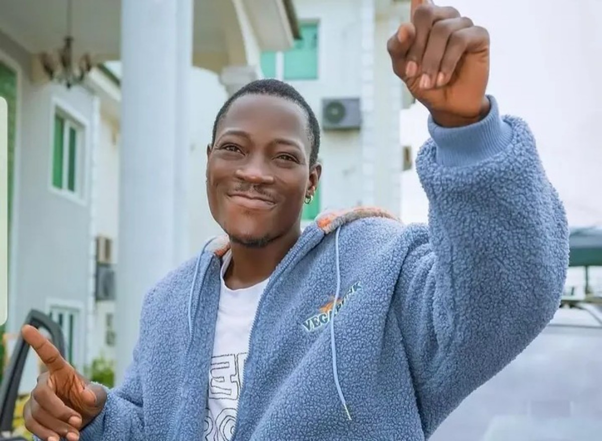 Smiling man in a light blue fleece jacket raises a clenched fist in the air outdoors, pointing with his other hand in a celebratory pose.