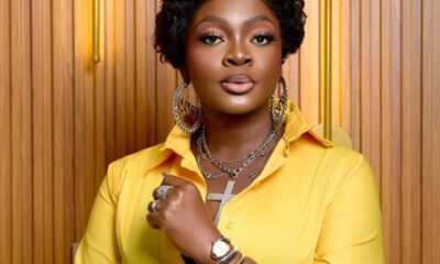 Portrait of a woman with short curly black hair wearing a bright yellow shirt and large silver jewelry, posed with arms crossed.