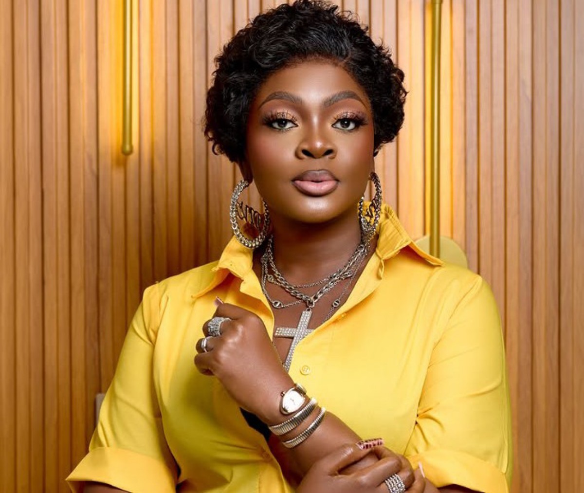 Portrait of a woman with short curly black hair wearing a bright yellow shirt and large silver jewelry, posed with arms crossed.