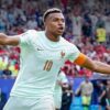 France captain wearing a light blue jersey with number 10, arms outstretched in celebration on the pitch, crowd in the stadium behind him.