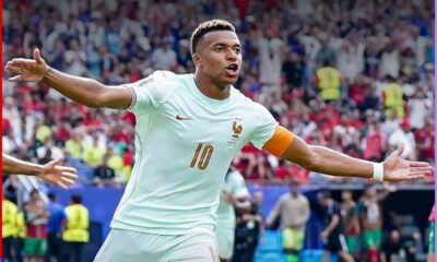 France captain wearing a light blue jersey with number 10, arms outstretched in celebration on the pitch, crowd in the stadium behind him.