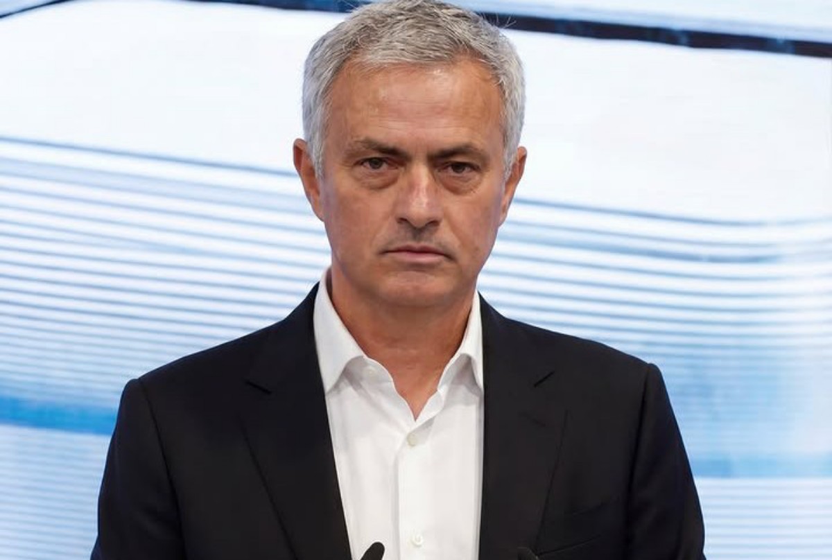 A man in a dark suit and white shirt speaks at a podium against a light, wavy backdrop.
