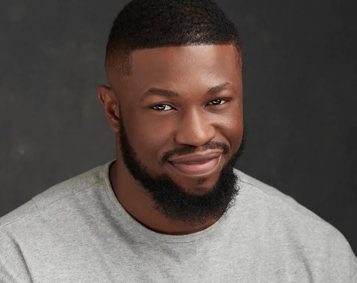 Close-up portrait of a smiling man with a short fade haircut and trimmed beard, wearing a gray shirt against a dark background.