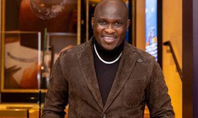 Smiling man in a brown suede blazer and black turtleneck, wearing a pearl necklace, indoors.