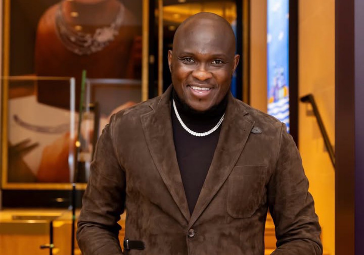 Smiling man in a brown suede blazer and black turtleneck, wearing a pearl necklace, indoors.