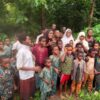 A large group of children and a few adults stand together in a lush, green outdoor area, looking at the camera.