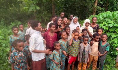 A large group of children and a few adults stand together in a lush, green outdoor area, looking at the camera.