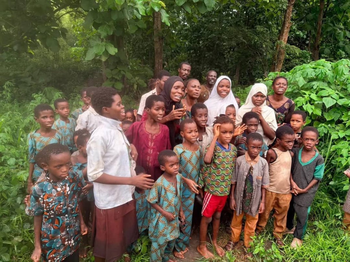 A large group of children and a few adults stand together in a lush, green outdoor area, looking at the camera.