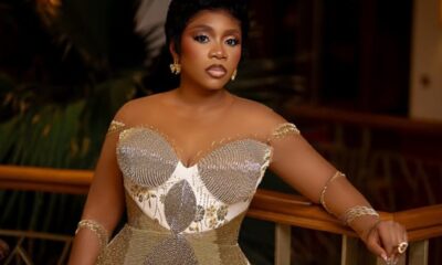 Woman in a gold, beaded off-the-shoulder gown posing by a railing, with short haircut and gold jewelry.