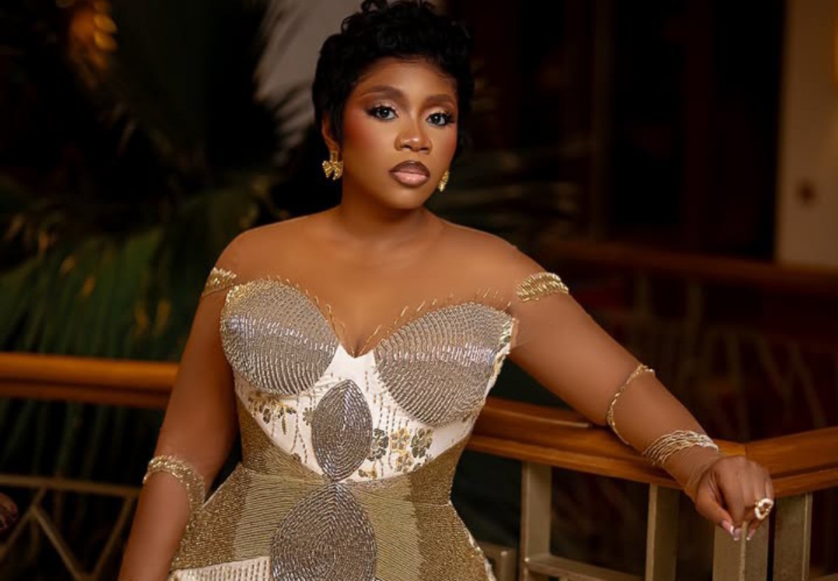Woman in a gold, beaded off-the-shoulder gown posing by a railing, with short haircut and gold jewelry.
