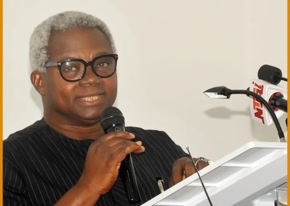 Speaker with gray hair and black glasses delivering a speech at a podium, holding a microphone.