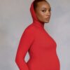 Pregnant woman in a red turtleneck bodysuit posing in profile against a gray backdrop
