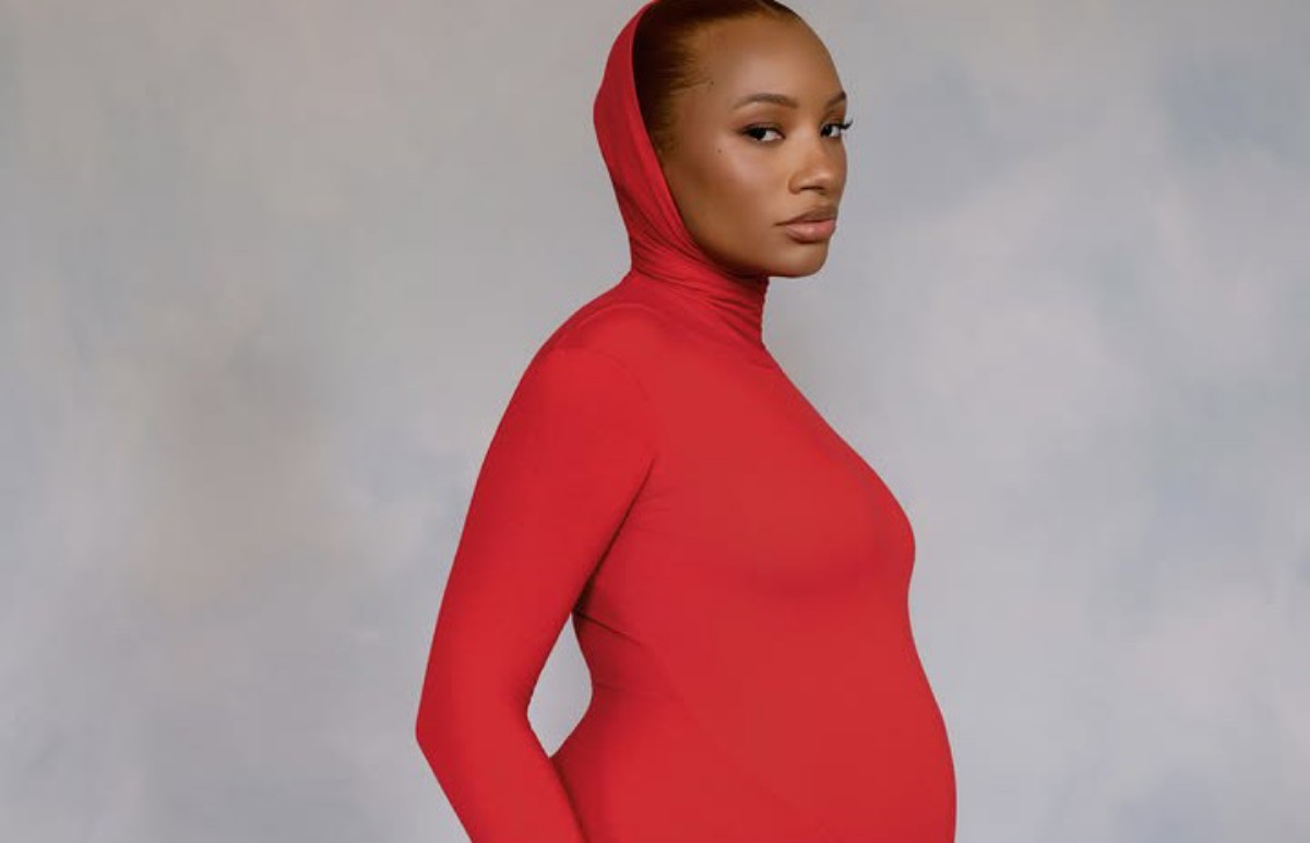 Pregnant woman in a red turtleneck bodysuit posing in profile against a gray backdrop