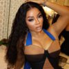 Portrait of a woman with long curly black hair wearing a blue halter dress, posing with arms raised in a vanity-lit room.