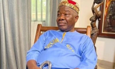 Older man in a blue traditional outfit and a gold crown, sitting in a wooden chair indoors, holding a cane.