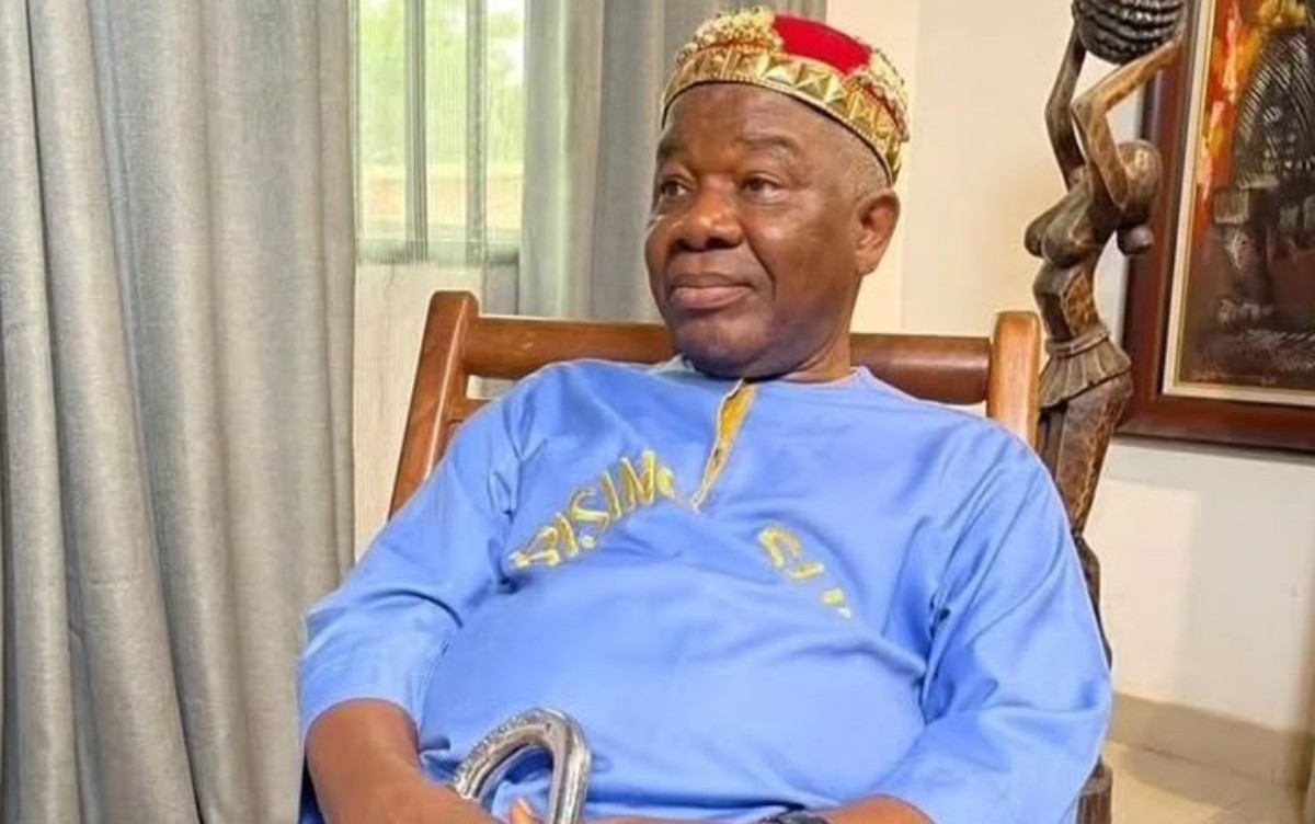 Older man in a blue traditional outfit and a gold crown, sitting in a wooden chair indoors, holding a cane.