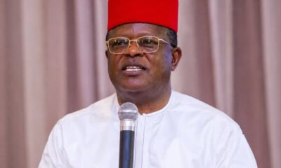 A man in a bright red fez and white traditional robe speaks into a handheld microphone.