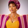 Portrait of a woman wearing a magenta gele headwrap, striped cape, and a yellow corset-style dress with gold jewelry.