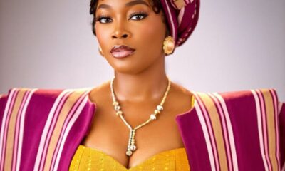 Portrait of a woman wearing a magenta gele headwrap, striped cape, and a yellow corset-style dress with gold jewelry.