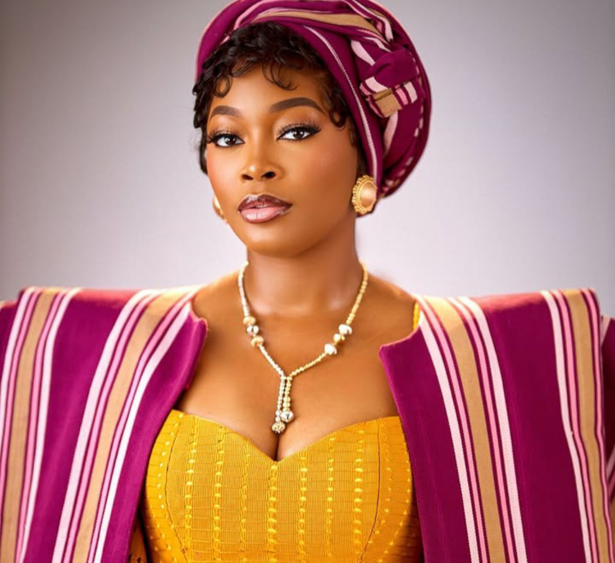 Portrait of a woman wearing a magenta gele headwrap, striped cape, and a yellow corset-style dress with gold jewelry.