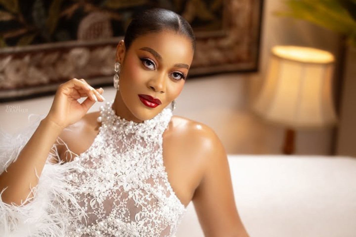 Elegant woman wearing a white lace high-neck dress with pearl embellishments and bold red lipstick, posing on a sofa.