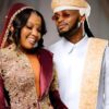 Couple in ornate traditional outfits smiling together at a celebration; the woman wears burgundy with gold embroidery, the man wears white with gold sash and headwrap