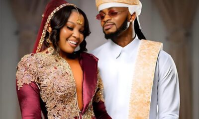 Couple in ornate traditional outfits smiling together at a celebration; the woman wears burgundy with gold embroidery, the man wears white with gold sash and headwrap