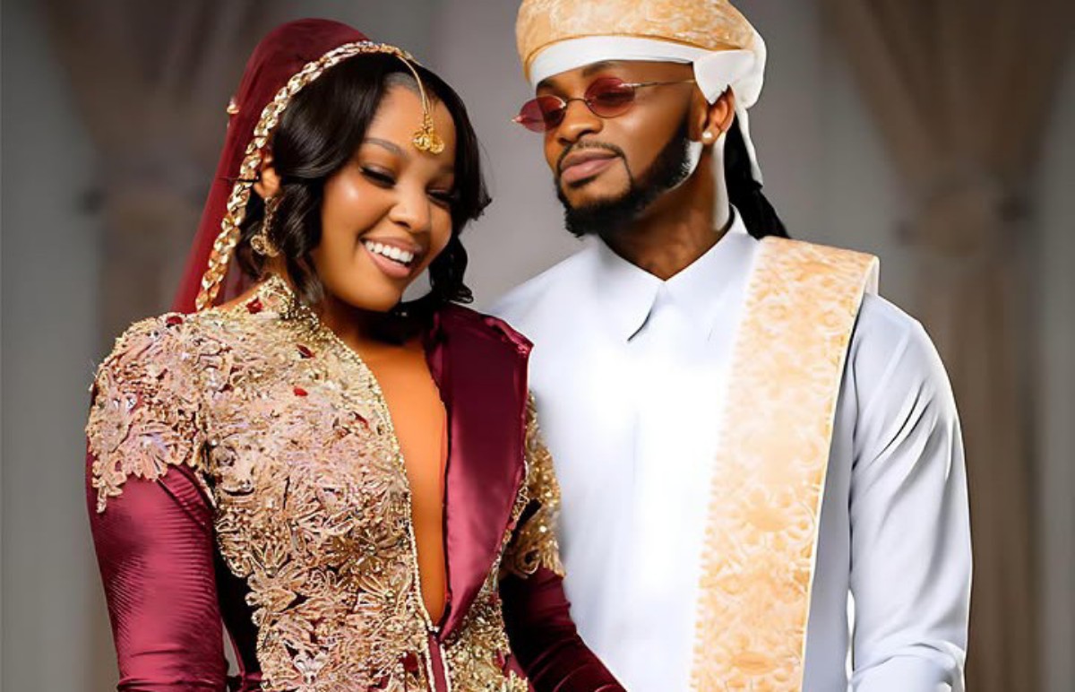 Couple in ornate traditional outfits smiling together at a celebration; the woman wears burgundy with gold embroidery, the man wears white with gold sash and headwrap