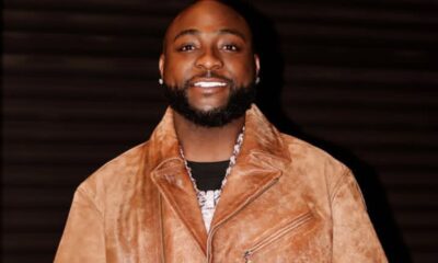 Smiling man with a trimmed beard wearing a tan suede jacket and silver necklace against a dark background.