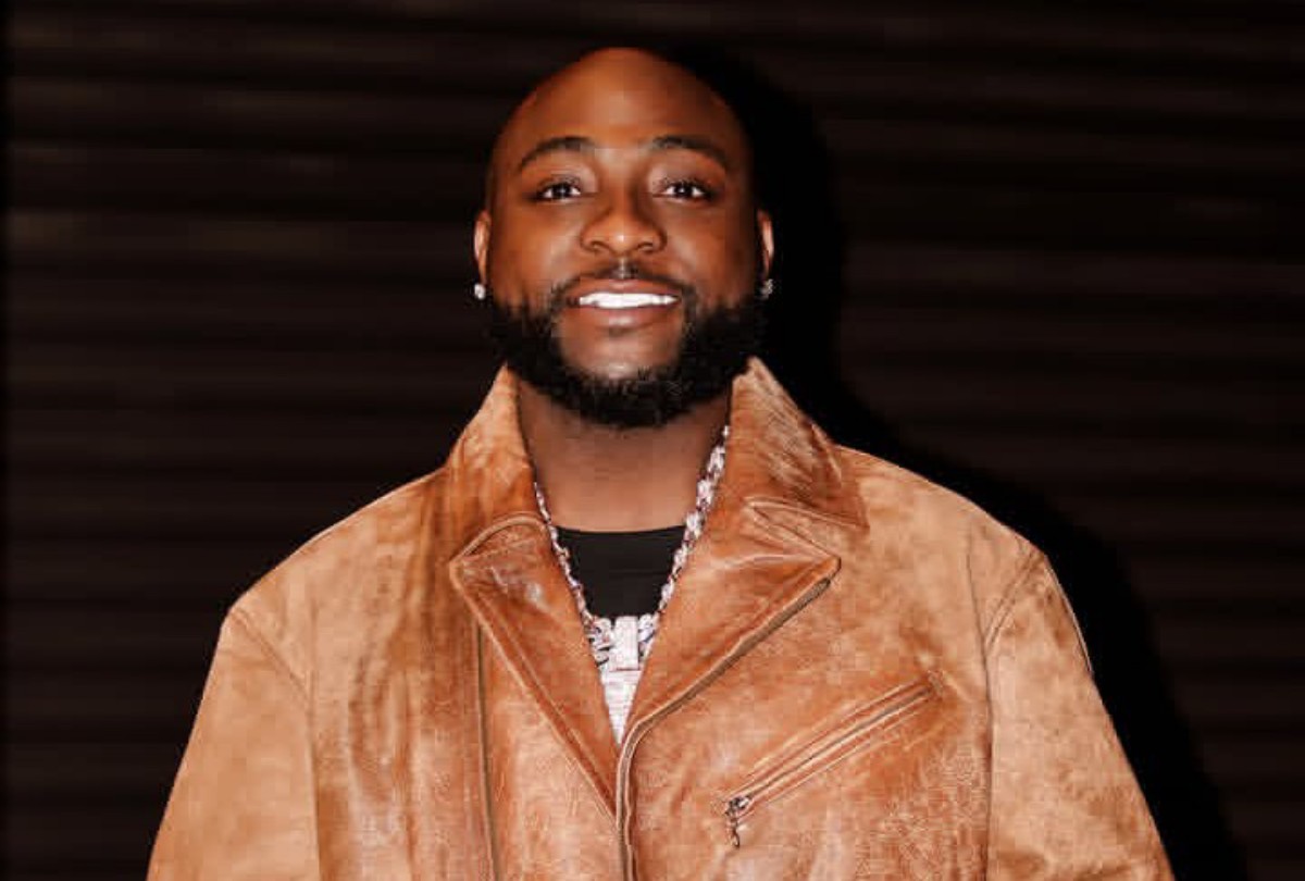 Smiling man with a trimmed beard wearing a tan suede jacket and silver necklace against a dark background.