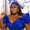 Woman in a royal blue dress with a ruched bodice and large ruffled sleeves, wearing a feathered blue headpiece and pearls, posing against light curtains.