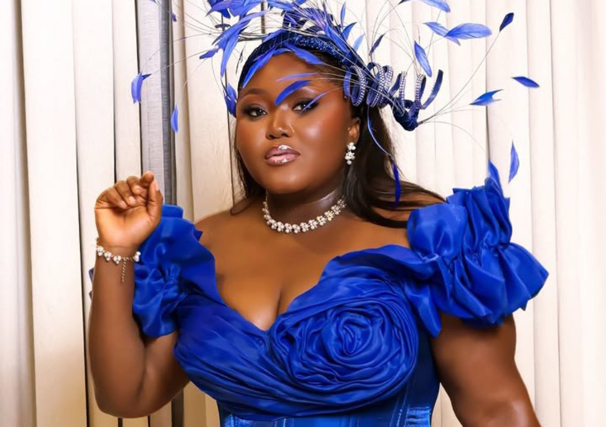 Woman in a royal blue dress with a ruched bodice and large ruffled sleeves, wearing a feathered blue headpiece and pearls, posing against light curtains.