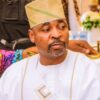 Man in white traditional attire and beige cap seated at a formal event, looking to his left.