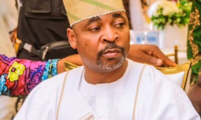 Man in white traditional attire and beige cap seated at a formal event, looking to his left.