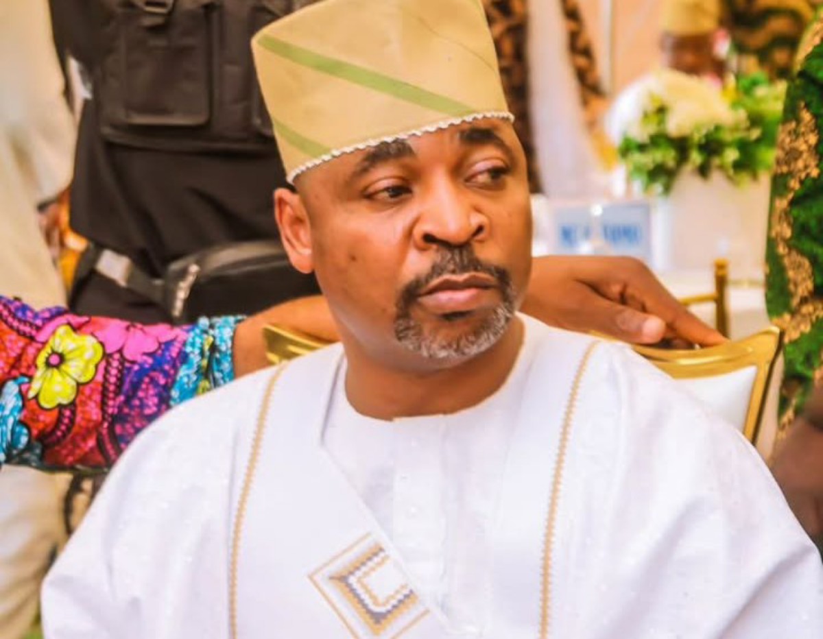 Man in white traditional attire and beige cap seated at a formal event, looking to his left.