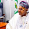 Man in white traditional attire and patterned cap sits at a desk in an office, with stacked books and a small Nigerian flag nearby.