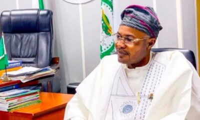 Man in white traditional attire and patterned cap sits at a desk in an office, with stacked books and a small Nigerian flag nearby.