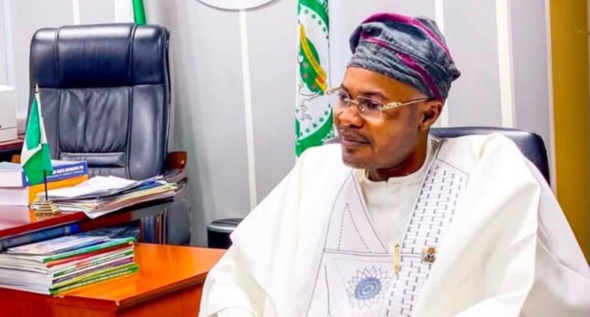 Man in white traditional attire and patterned cap sits at a desk in an office, with stacked books and a small Nigerian flag nearby.